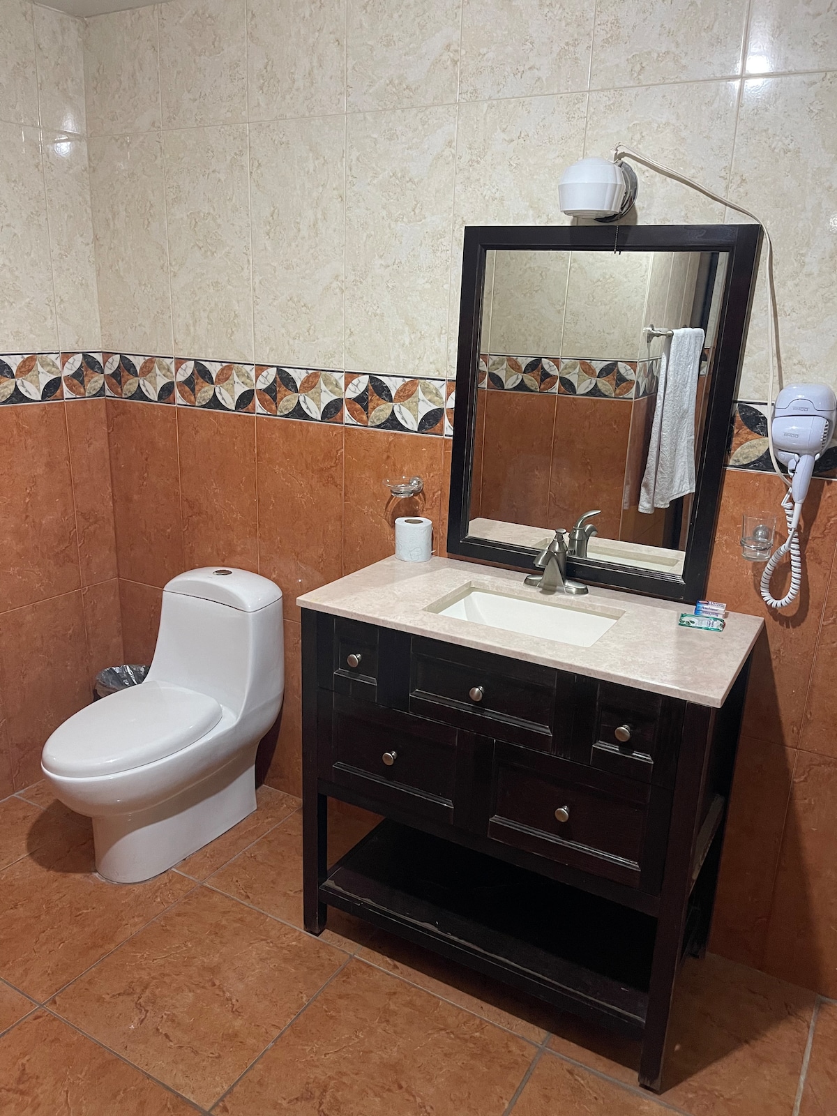 A functional bathroom features tiled walls with a decorative border. A white toilet is positioned beside a dark wooden vanity, which includes a sink and storage drawers. A large mirror above the sink reflects the space, and a hairdryer is attached to the wall.