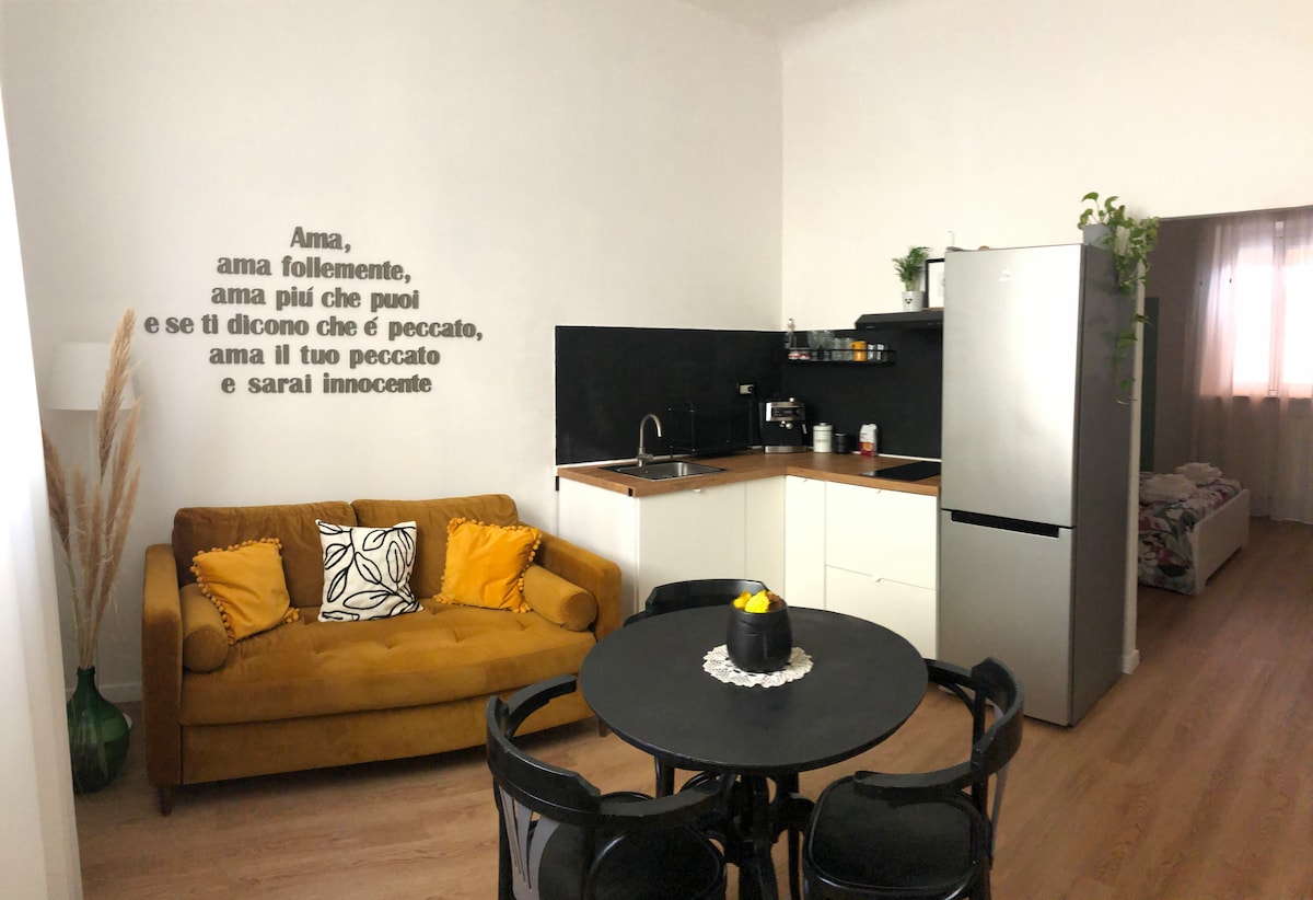 An inviting living space features a cozy brown sofa adorned with yellow pillows. A circular dining table with black chairs sits nearby, complemented by a modern corner kitchen with sleek cabinetry and stainless steel appliances. Natural light filters in through a window, enhancing the atmosphere.