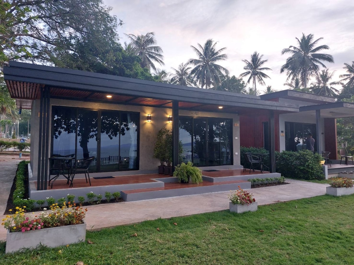 A modern resort structure features large glass windows that provide views of the sea. The exterior is surrounded by lush greenery and includes a small garden area filled with colorful flowers. Chairs and tables are positioned for outdoor seating on the patio.
