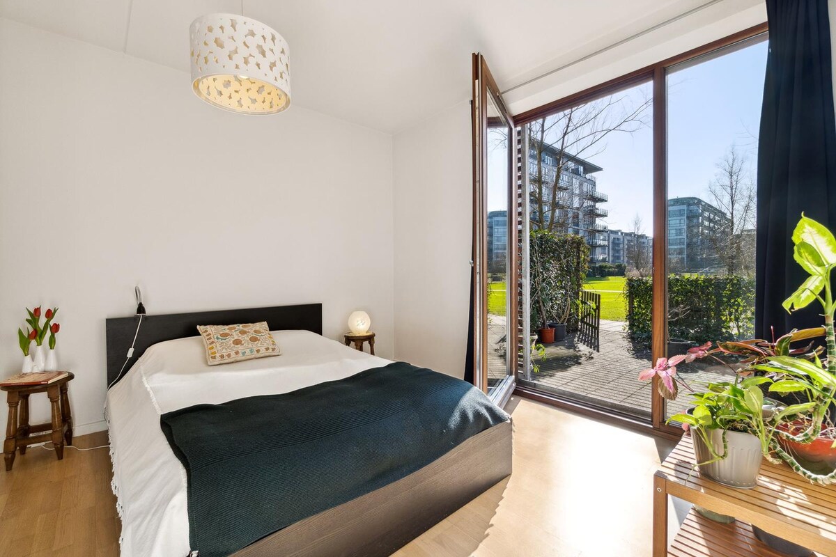 A bedroom features a spacious bed with a dark throw and decorative pillow. Natural light floods in through large glass doors that open to a garden view. A small wooden table holds a vase of flowers, while potted plants bring a touch of greenery to the space.