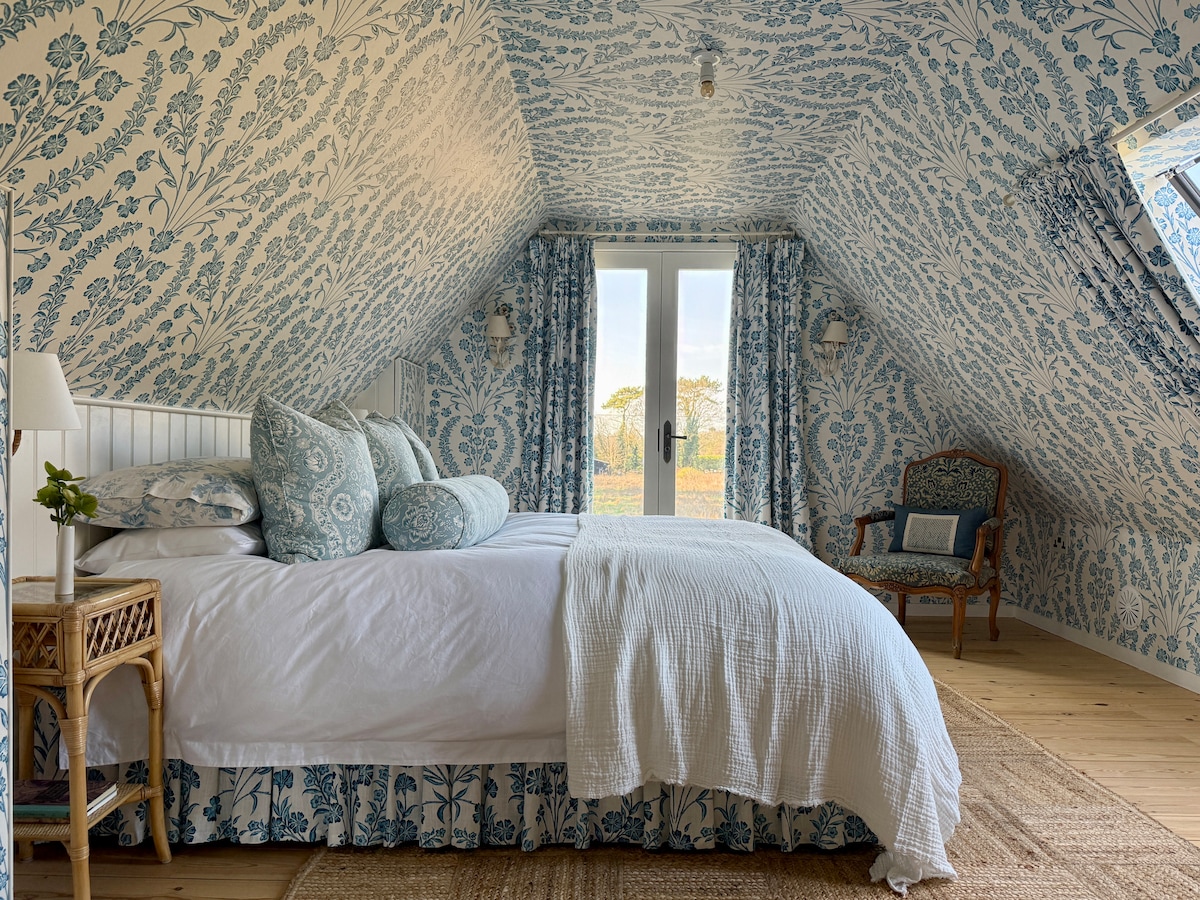A room features sloped ceilings adorned with blue floral wallpaper. A spacious bed is layered with soft linens and multiple pillows. Natural light enters through double doors leading to outdoor views. A cozy chair is positioned beside the window, complemented by a decorative side table.