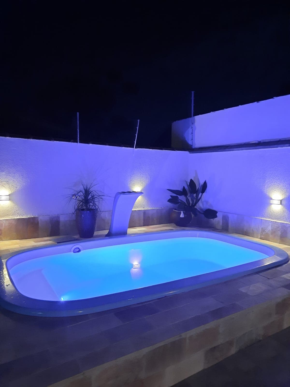 A serene swimming pool is illuminated with soft blue lights, creating a calming atmosphere. The pool features a smooth edge with a flowing water feature, complemented by potted plants in the background. The surrounding wall provides an enclosed feeling for relaxation.