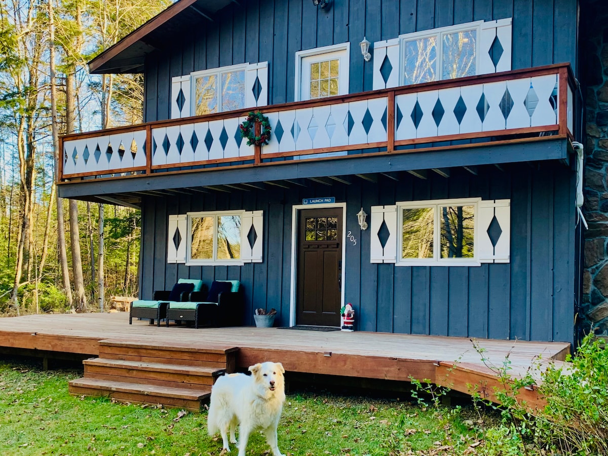 The exterior of the duplex home features a dark wood facade complemented by white diamond accents. A spacious deck is lined with seating options, and a friendly dog stands in front. Large windows allow natural light to fill the interior, enhancing the inviting atmosphere.