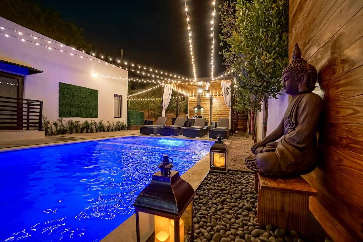 A vibrant blue pool is illuminated by soft lighting strung overhead, creating a serene outdoor setting. Comfortable lounge chairs are arranged nearby, while a decorative lantern and a stone statue add to the tranquil atmosphere. Greenery lines the area, enhancing the secluded feel.