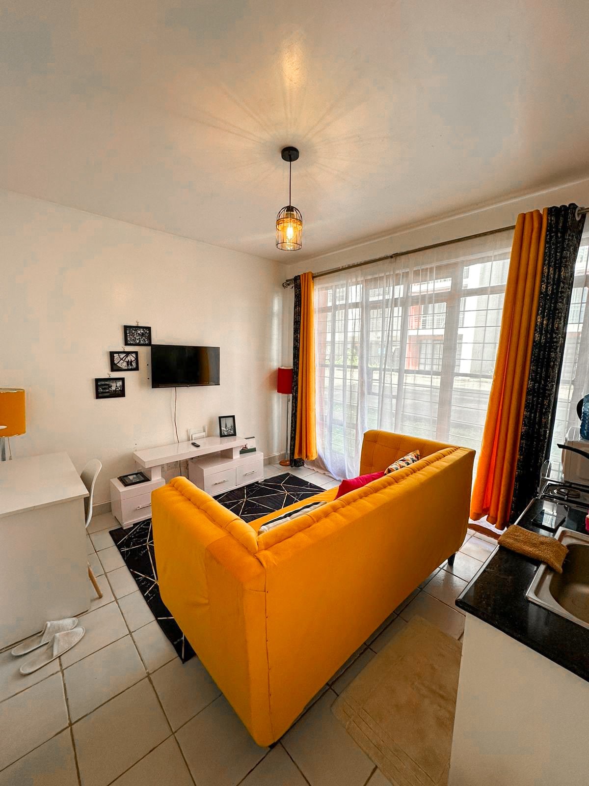 A bright living area features a yellow sofa that adds a pop of color. A flat-screen television is mounted on the wall, with a white media console below. Natural light filters through the large window, complemented by orange and black curtains. A workspace with a chair is situated nearby.