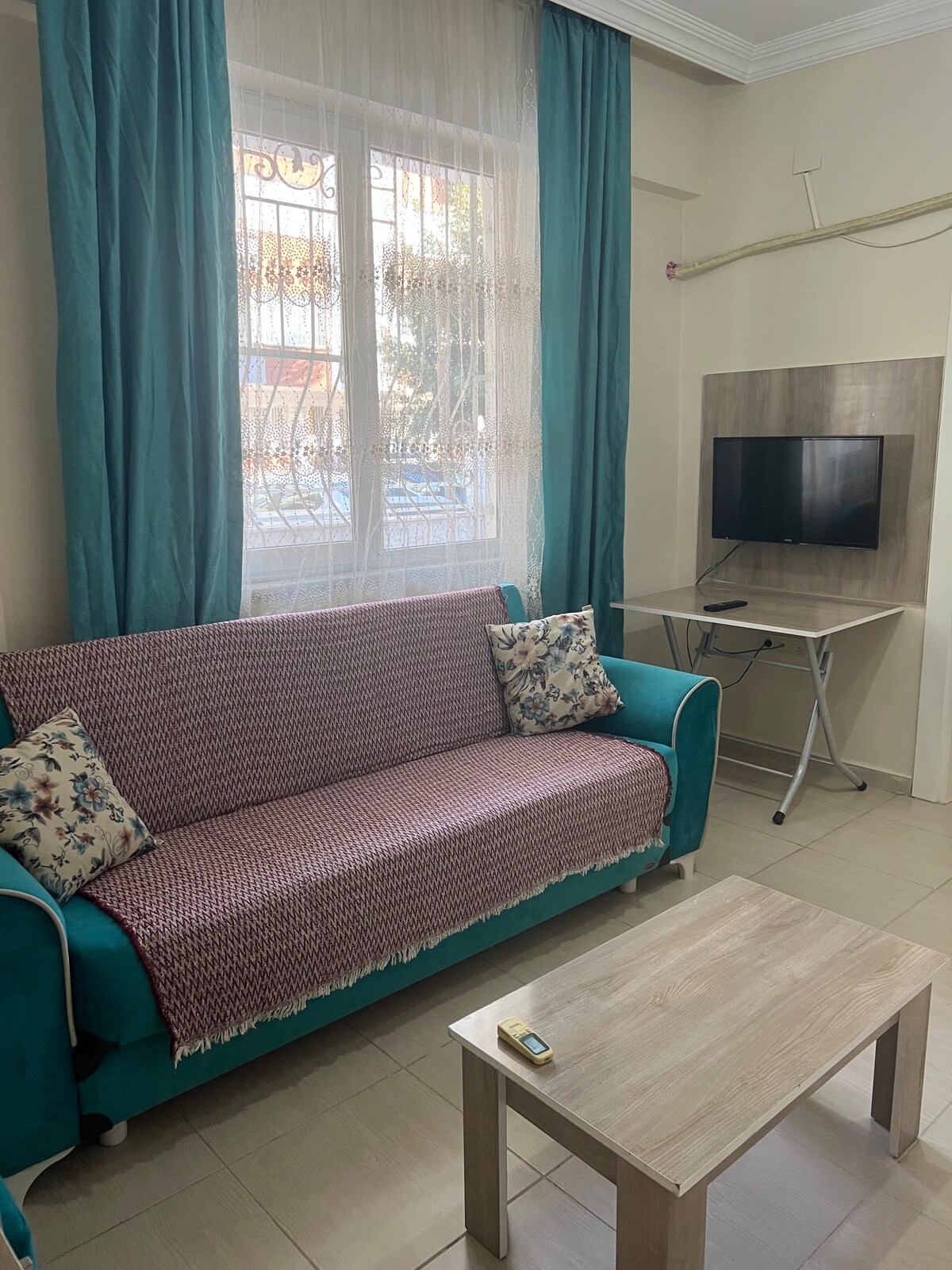 A cozy living area is presented with a teal sofa adorned with patterned cushions. A soft throw blanket drapes over the back. A light wooden coffee table sits in front, and a wall-mounted television is visible nearby. Natural light enters through sheer curtains at the window.