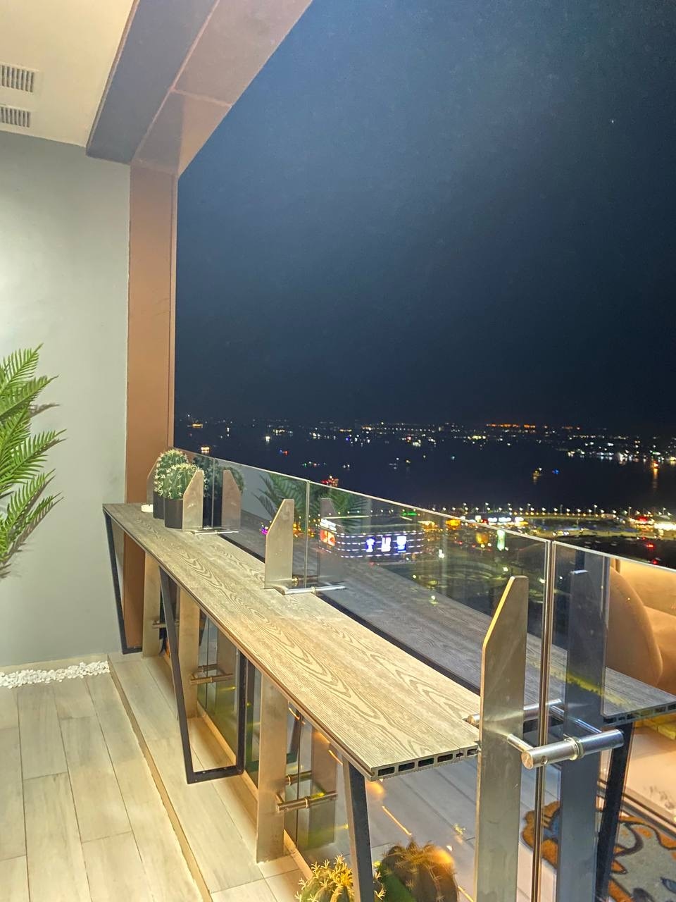 A balcony view showcases a modern wooden bar table with elegant metallic supports, overlooking a vibrant city landscape. Night lights illuminate the surroundings, enhancing the scene. A potted plant adds a touch of greenery, while the expansive skyline stretches in the background.