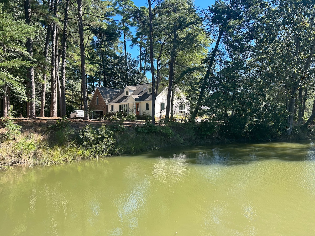 A charming retreat is positioned beside a serene body of water, surrounded by tall trees. The house features a mix of traditional and modern architecture, with large windows reflecting the natural beauty of the setting.