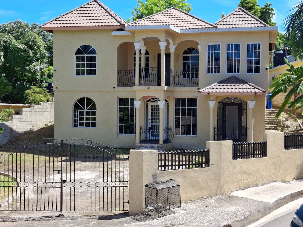 A two-story villa features a light-colored exterior with arched windows and decorative columns. The gated entrance is visible, leading to a spacious front yard. Lush greenery surrounds the property, enhancing the inviting atmosphere of the home.