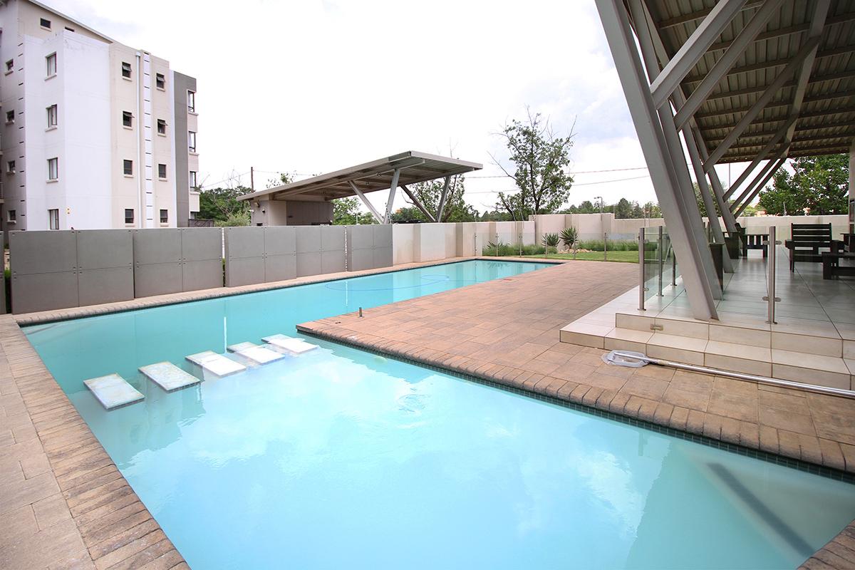 A modern swimming pool is surrounded by a tiled area, featuring a series of stepping stones leading across the water. Shade is provided by a sleek roof structure. Nearby, seating arrangements are visible against a backdrop of greenery and well-maintained landscaping.