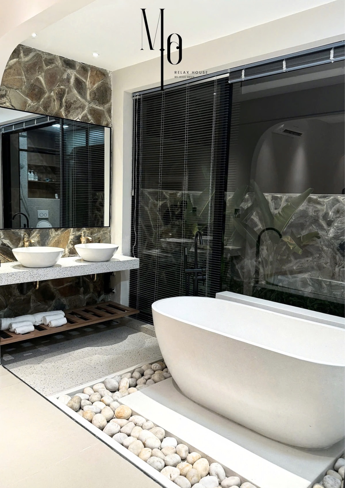 A modern bathroom features a freestanding tub positioned on smooth pebbles, with two sinks situated on a textured countertop. A large mirror reflects the natural stone wall, while floor-to-ceiling windows allow light to fill the space, offering a serene connection to the outdoors.
