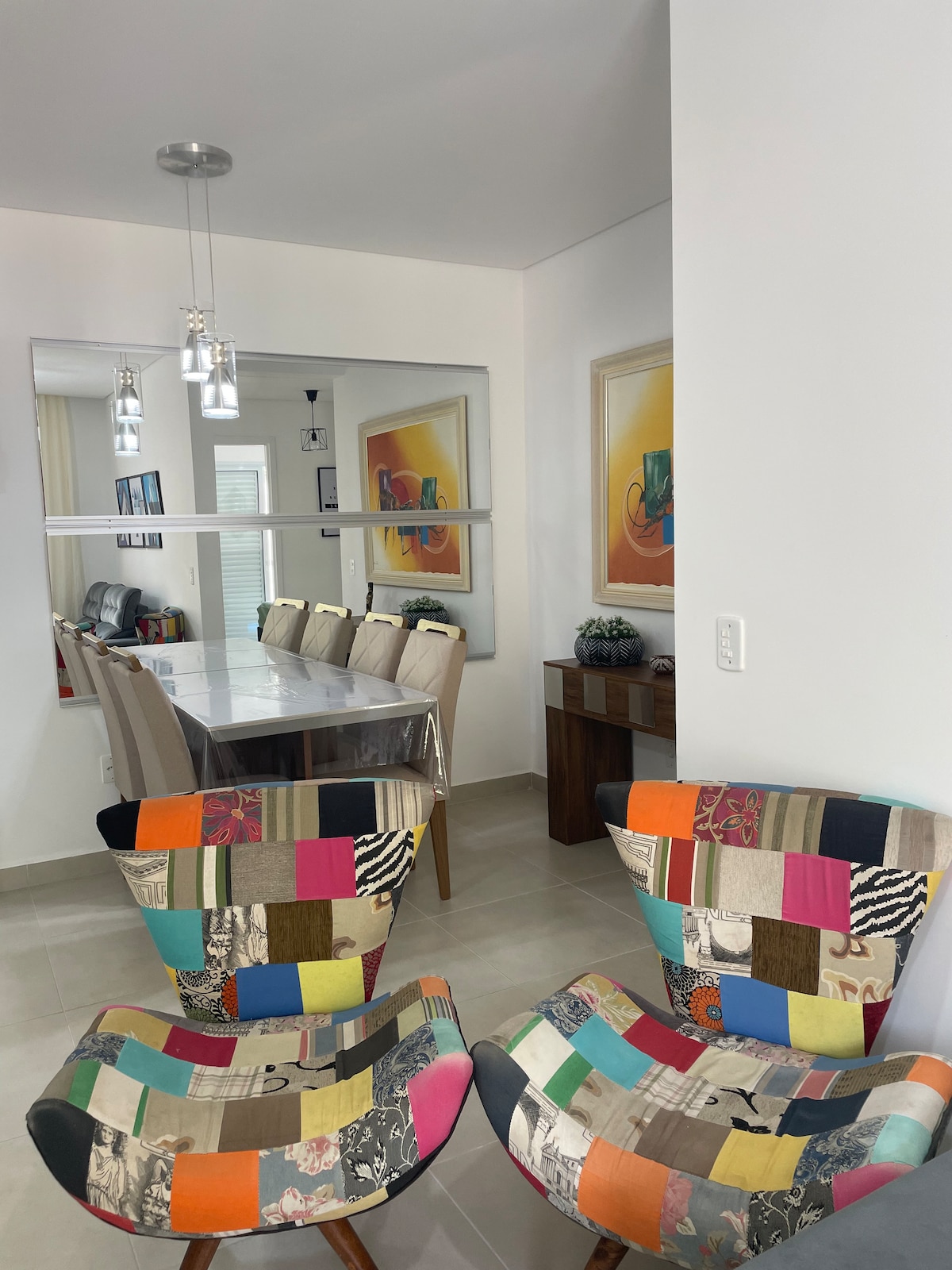 Two patchwork chairs with a mix of colorful fabrics are positioned in the foreground. A dining table is visible in the background, surrounded by light-colored upholstered chairs. Decorative artworks adorn the walls, and a console table is seen against the wall, enhancing the overall contemporary décor.
