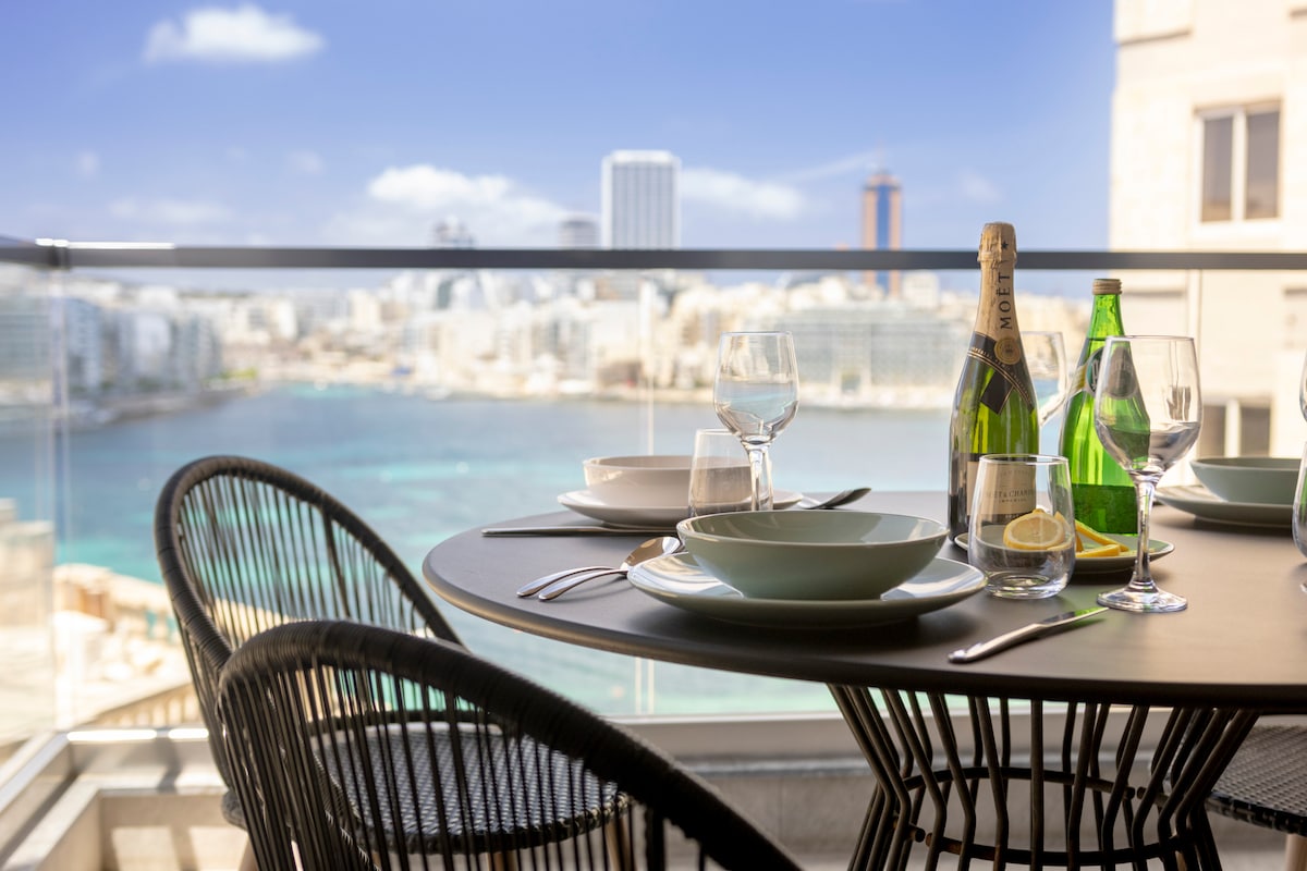 A dining table is set for four on a balcony overlooking the vibrant blue waters of St. Julian's Bay. Glassware, a bottle of champagne, and plates are arranged neatly, inviting guests to enjoy a meal with a stunning backdrop of the city skyline.