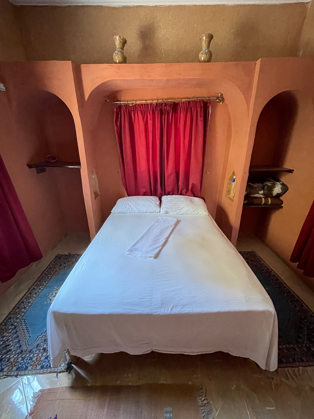 A cozy sleeping area features a spacious bed dressed in white linens and positioned beneath a small archway. Rich red curtains frame the bed, while decorative items are placed on the shelves nearby. A colorful rug adds warmth to the wooden floor.