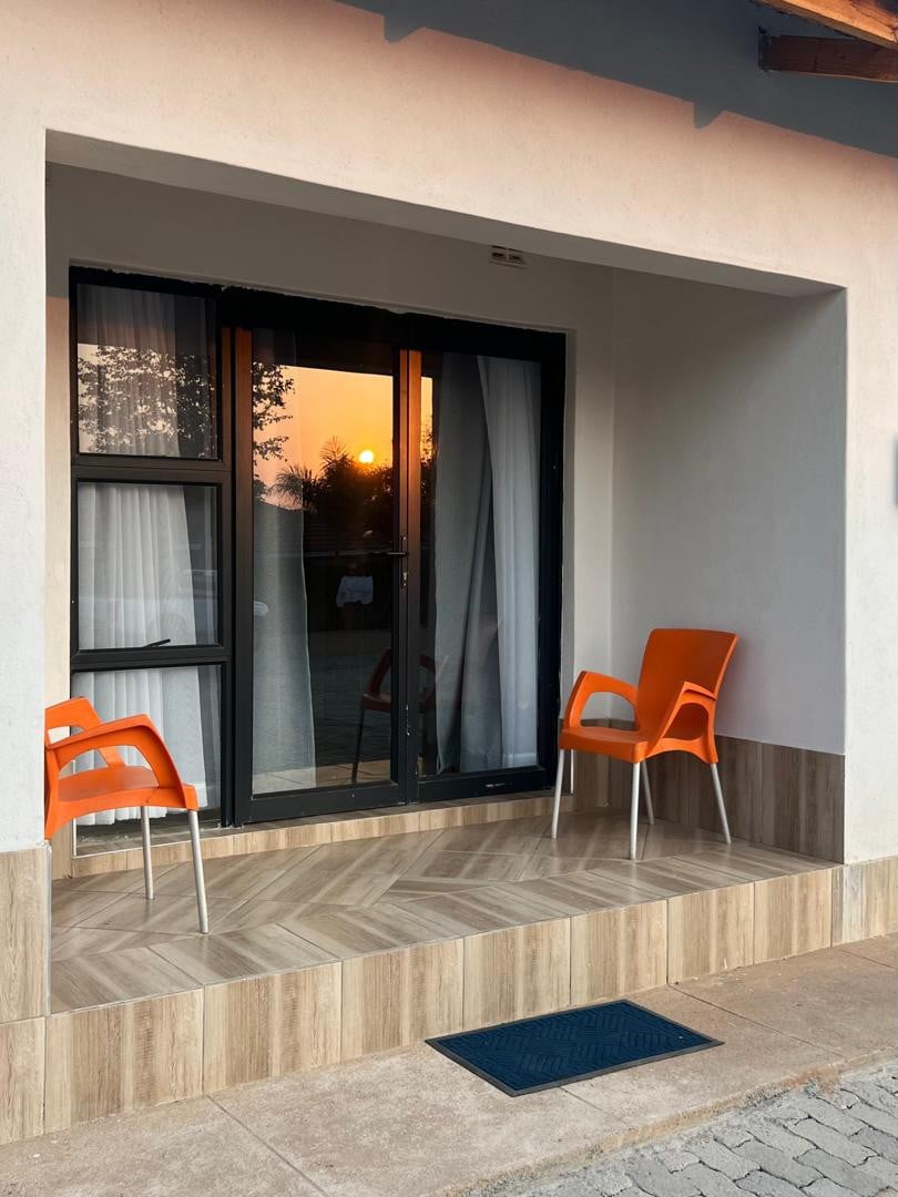 An outdoor patio area features a sleek entrance with floor-to-ceiling glass doors. Two bright orange chairs are positioned on a tiled terrace, which is bordered by light-colored walls. A view of the sunset is visible in the background, enhancing the tranquil setting.
