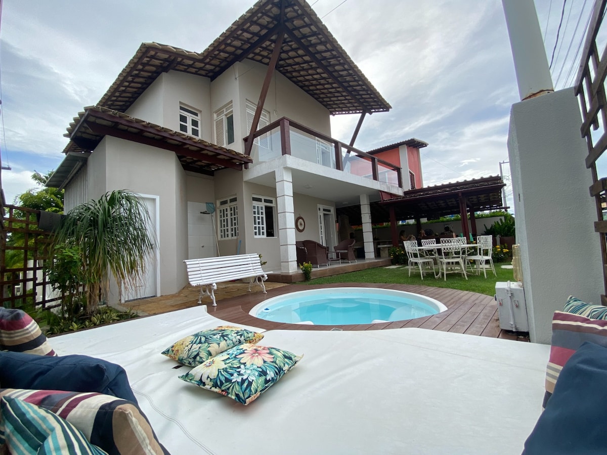 The exterior showcases a two-story house with a contrasting roof design. A small pool is set within a wooden deck surrounded by greenery. Lounge seating with decorative cushions is positioned nearby, while a dining area is visible in the garden space.