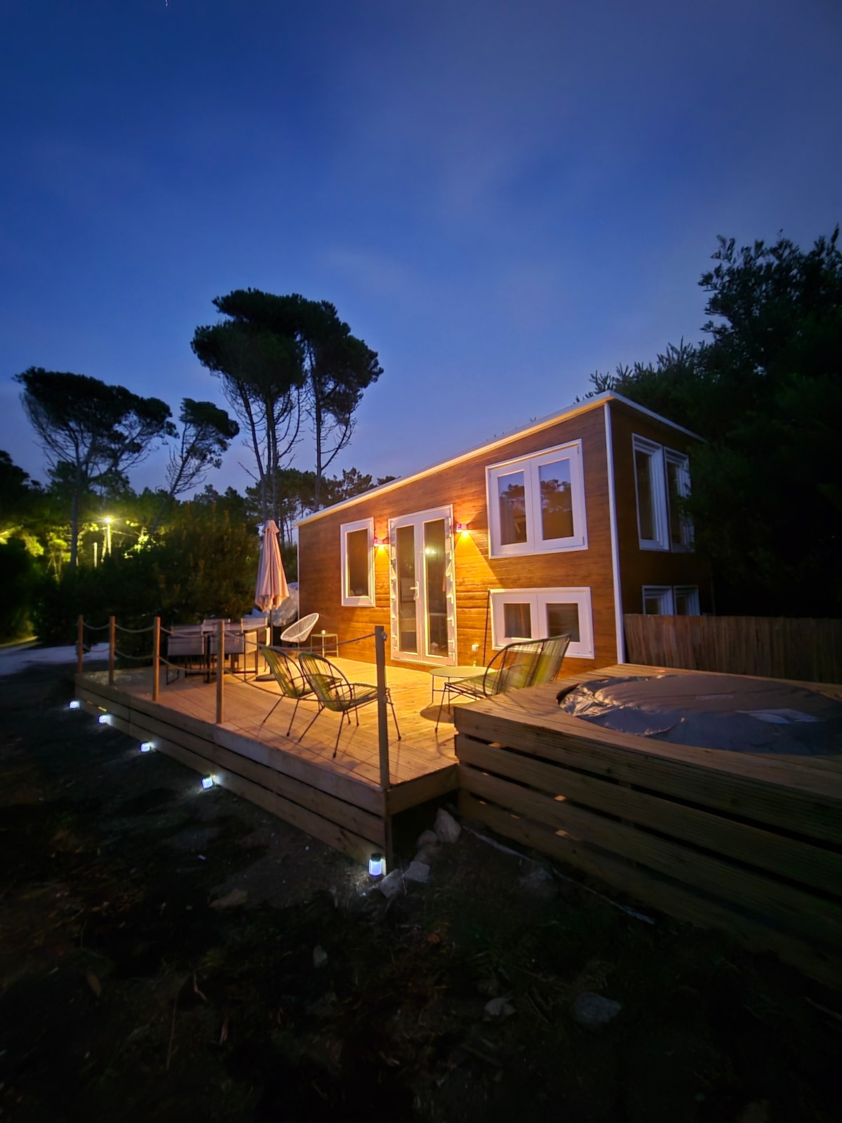 The tiny house is illuminated at dusk, showcasing large windows and a welcoming wooden deck. Outdoor seating is arranged with a table and chairs, while a jacuzzi is positioned nearby. Tall trees surround the area, enhancing the connection to nature.