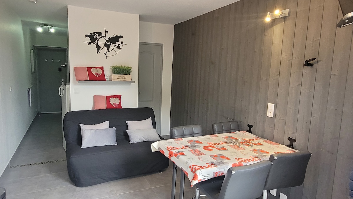 A cozy living space is depicted, featuring a black sofa bed with colorful cushions. A rectangular dining table surrounded by four grey chairs occupies the center. The wall is adorned with wooden panels, and soft lighting highlights the area, creating a comfortable atmosphere.