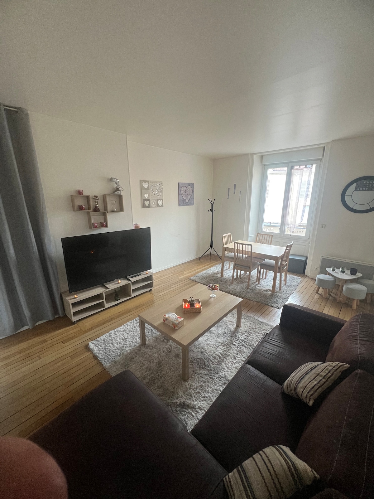 A cozy living area features a dark sofa facing a television stand. A light wood dining table is surrounded by four chairs. Soft textures are present with a rug beneath the table, and natural light enters through a window with sheer curtains.