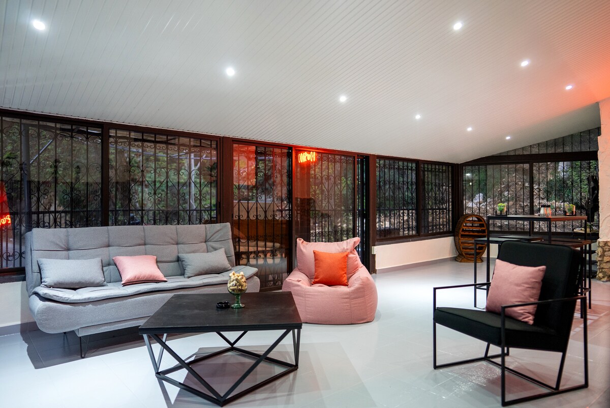A modern lounge area features large windows surrounded by greenery, allowing natural light to fill the space. A soft gray couch and a vibrant pink bean bag provide comfortable seating, complemented by a dark coffee table. A black chair accompanies the seating arrangement, enhancing the inviting atmosphere.