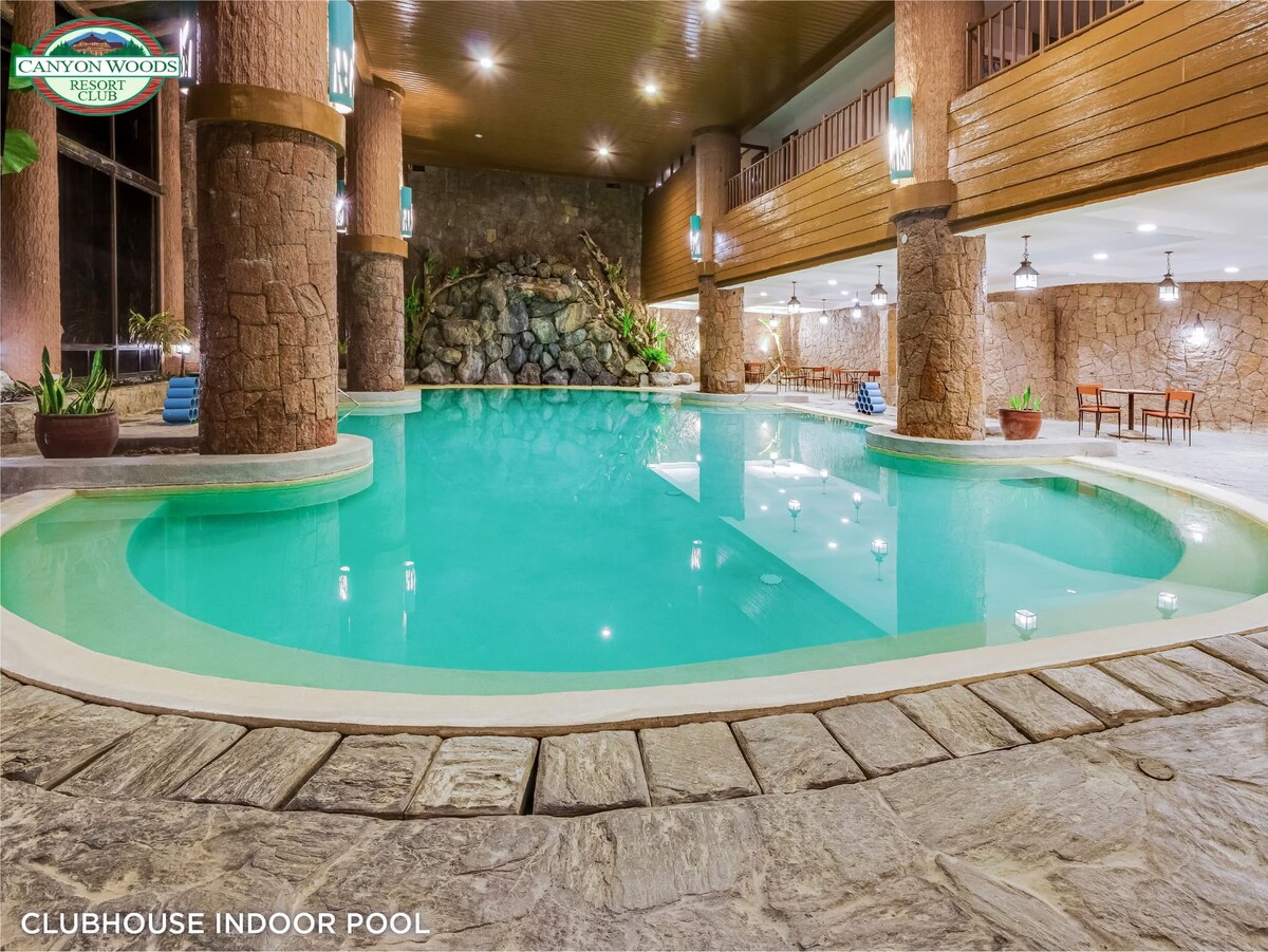 An indoor pool featuring calm turquoise water is surrounded by natural stone walls and complemented by wooden pillars. Lounge chairs are placed beside the pool, and lush greenery accents the space, creating a relaxing atmosphere for guests.
