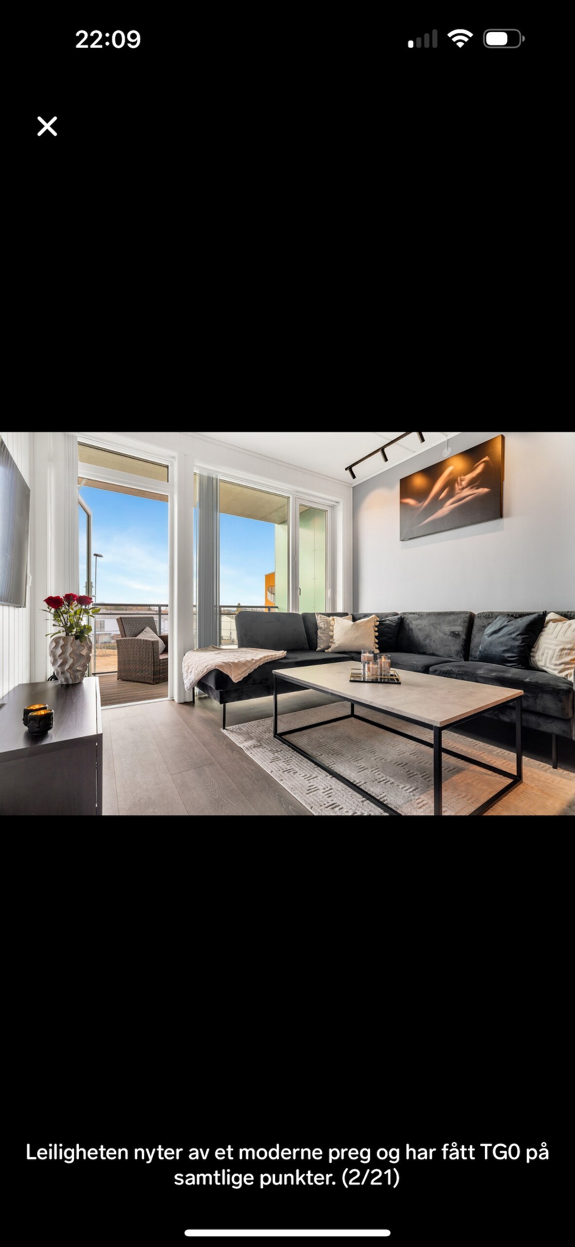 A modern living area features a sleek sofa positioned across from a low coffee table. Large glass doors open to a balcony, allowing natural light to fill the space. A piece of artwork adorns the wall, complementing the neutral color palette.