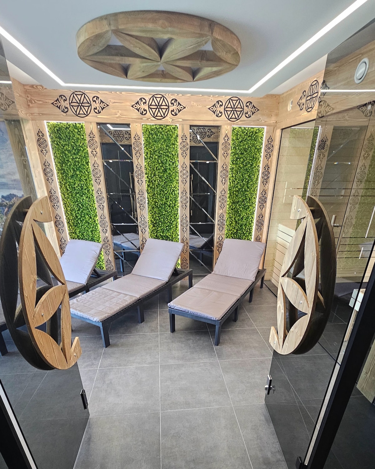 A serene relaxation area features comfortable lounge chairs arranged in a calming space. Green wall accents and light wood elements create a tranquil environment, while large glass doors provide access to the area.