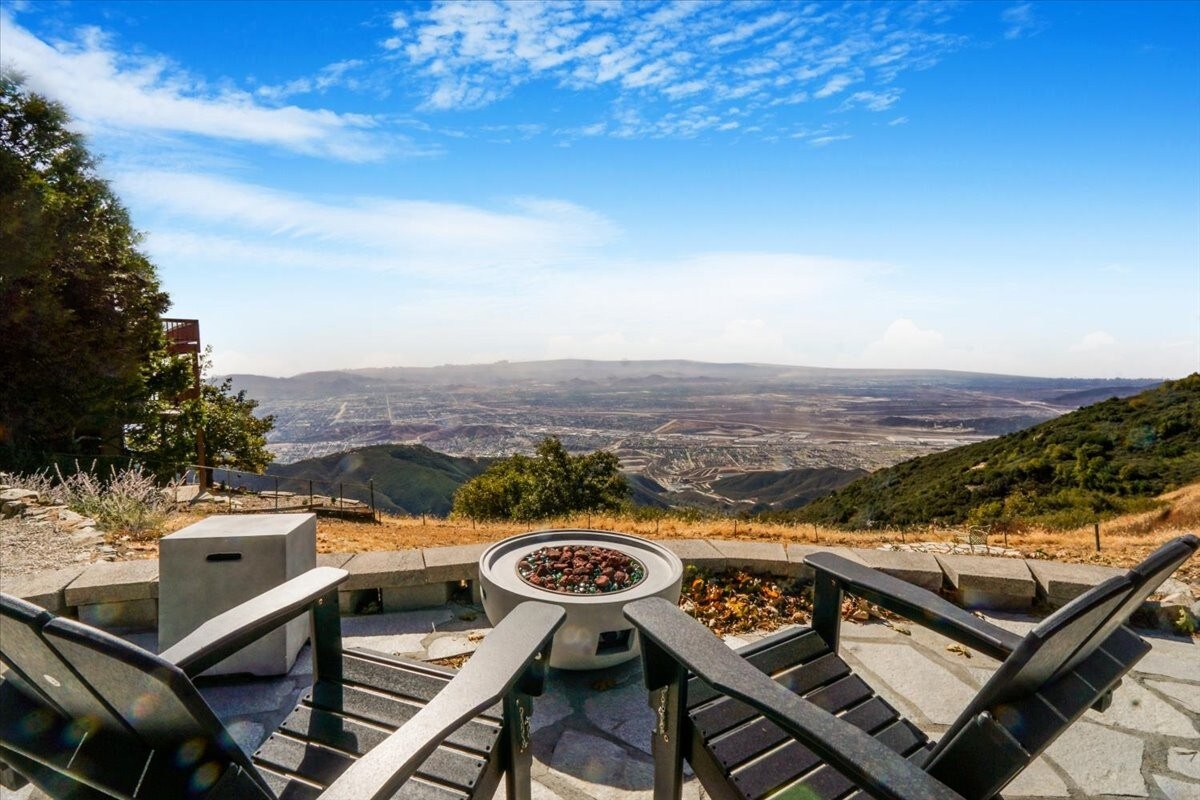 An outdoor seating area is shown, featuring multiple black lounge chairs arranged around a circular fire pit. A panoramic view of the valley below is visible, showcasing the landscape under a clear blue sky, with distant mountains adding depth to the scenery.