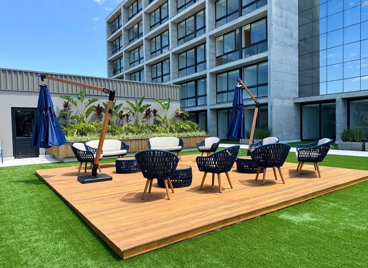 An outdoor relaxation area features a wooden deck surrounded by green grass. Multiple seating arrangements are made up of modern chairs and low tables, complemented by tall umbrellas. Lush greenery is visible in the background, enhancing the open space.
