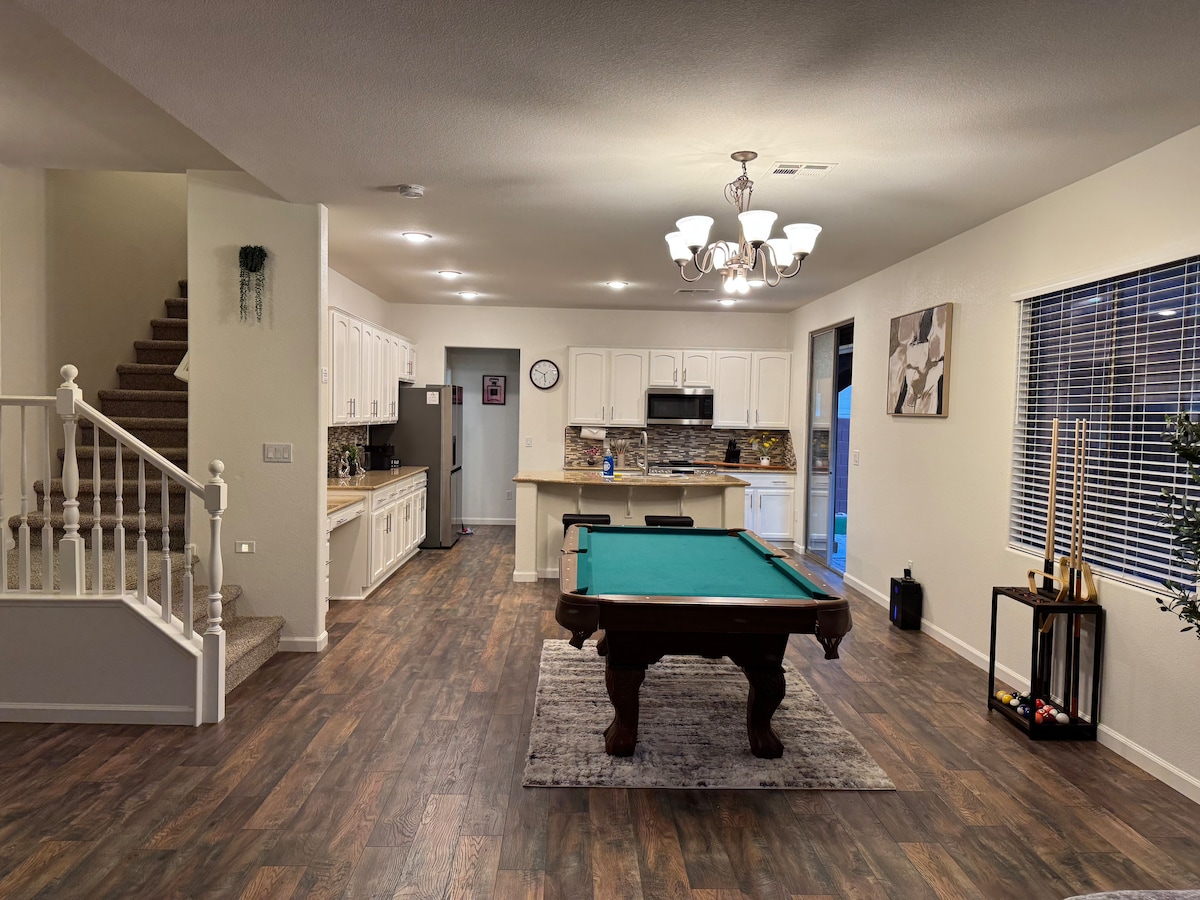 An open living space features a billiard table at the center, surrounded by hardwood flooring. A well-equipped kitchen is visible in the background, with white cabinetry and modern appliances. Natural light fills the area through large windows, and a staircase is situated to the left.