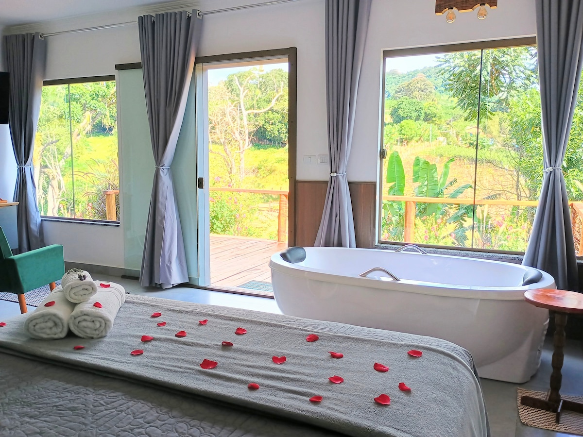 A spacious bedroom features large windows that allow for natural light and offer views of the surrounding greenery. A freestanding bathtub is positioned nearby, adorned with rose petals. A neatly made bed is complemented by rolled towels on top.