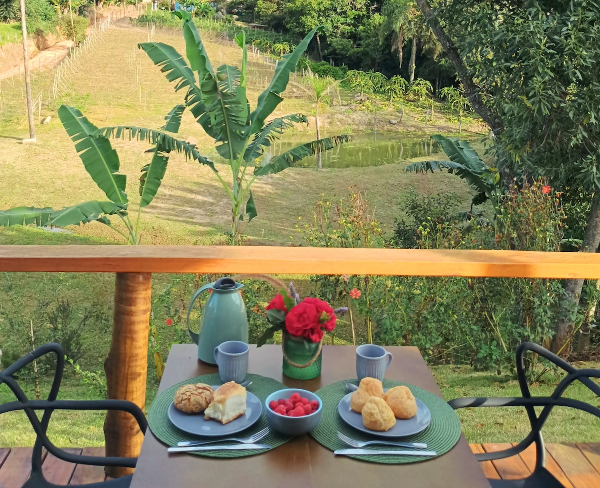 A dining table is set with plates of fresh pastries and fruit, alongside cups and a teapot. The view includes lush greenery, featuring banana plants and a vineyard in the background. The tranquil setting is enhanced by the natural landscape visible from the deck.