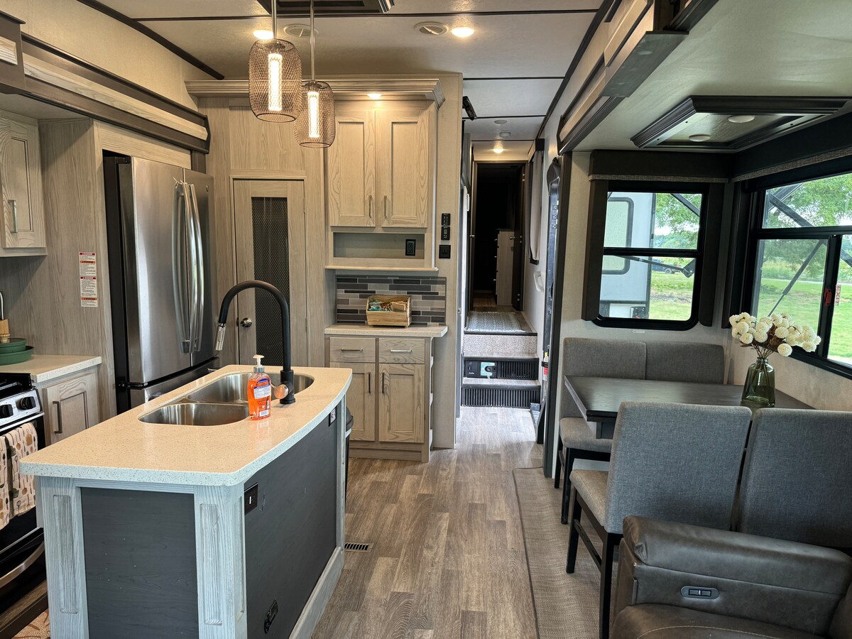 The interior of the RV features a modern kitchen with an island and bar seating. A dining area with a table and plush chairs is visible, along with a cozy seating area. Large windows allow natural light to illuminate the space.