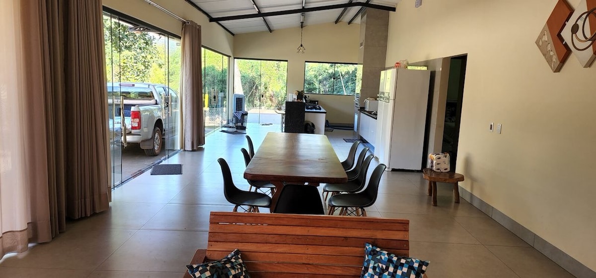 An open living space features a large wooden dining table accompanied by black chairs. Floor-to-ceiling windows offer views of the surrounding nature. A comfortable wooden bench and a well-equipped kitchen area are visible. Natural light brightens the room, enhancing the spacious atmosphere.