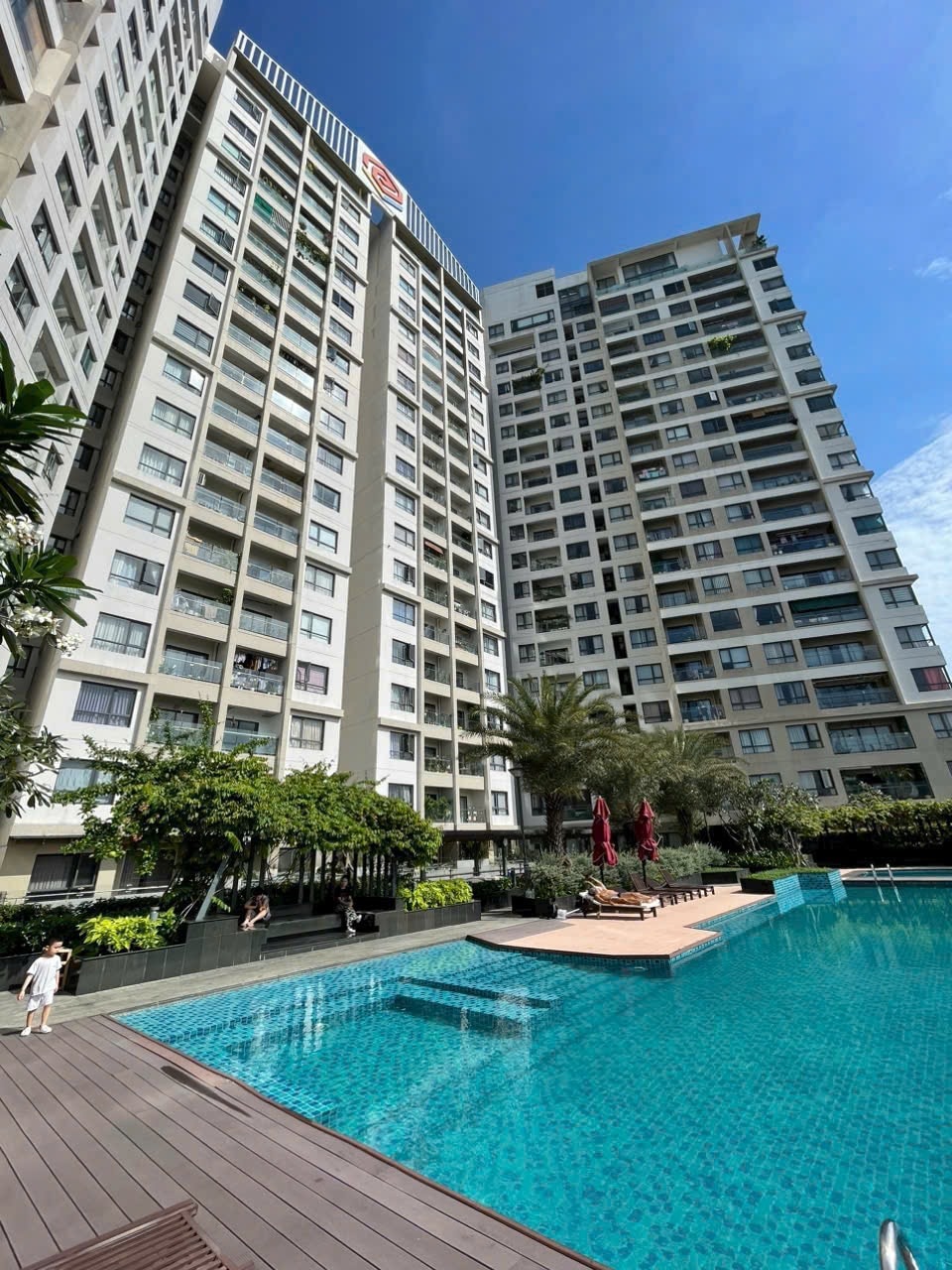 A modern high-rise building is depicted, surrounded by lush greenery. In the foreground, a clear blue swimming pool reflects the sunny sky, while sun loungers are positioned along the deck. The architecture features a mix of glass and concrete, enhancing its contemporary design.