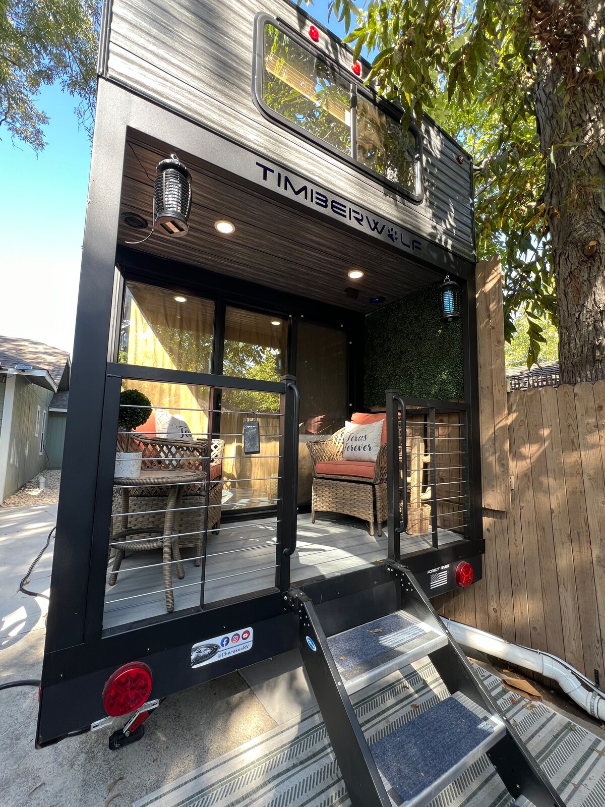 A stylish exterior of a tiny home is showcased, featuring a small set of stairs leading up to a welcoming entrance. The space is adorned with decorative seating and potted plants, providing a charming and inviting atmosphere.