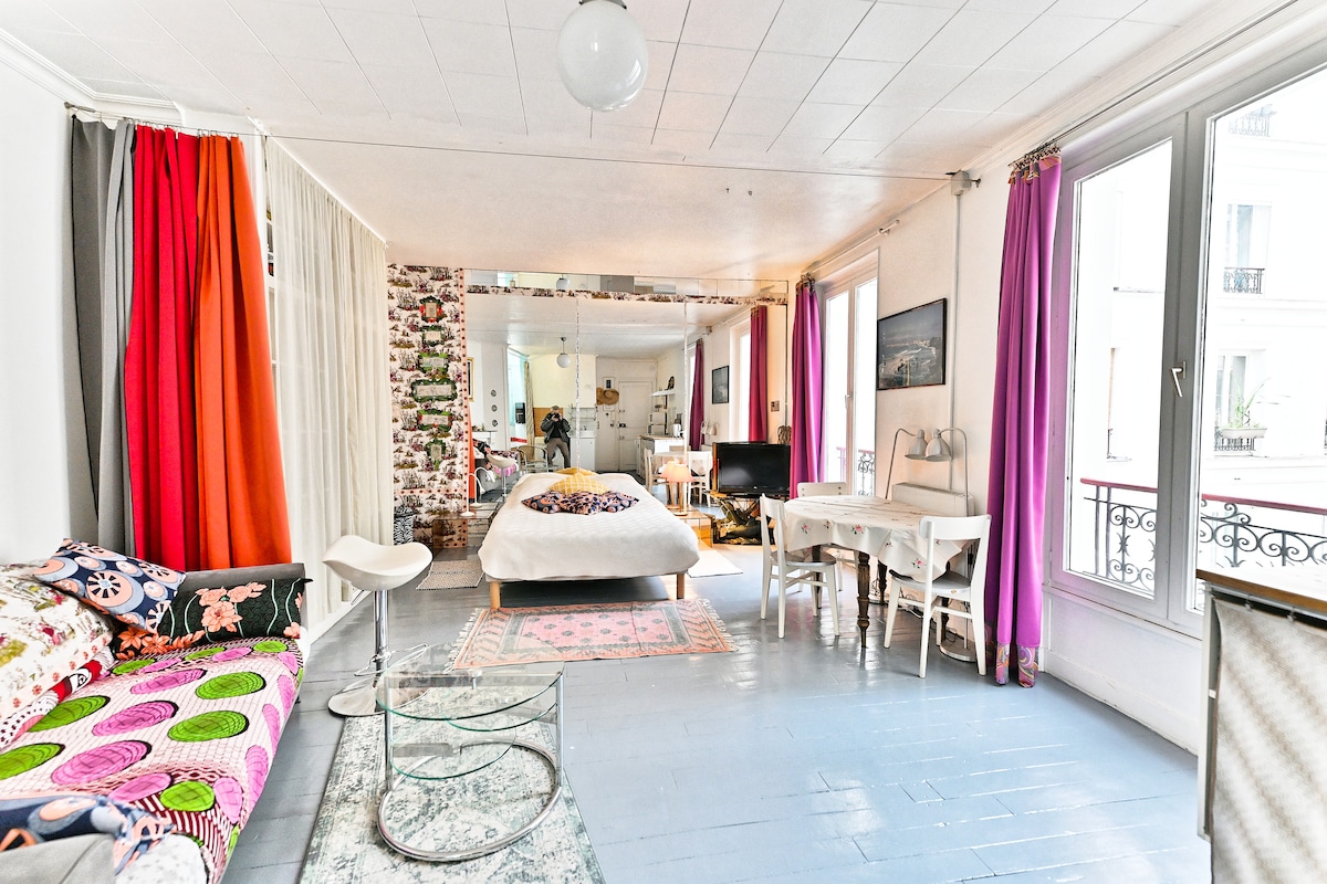 A spacious studio features large windows allowing natural light to fill the room. A comfortable double bed is centered, with vibrant curtains in various shades adding color. A cozy seating area includes a colorful sofa, while a small desk and television are also visible.