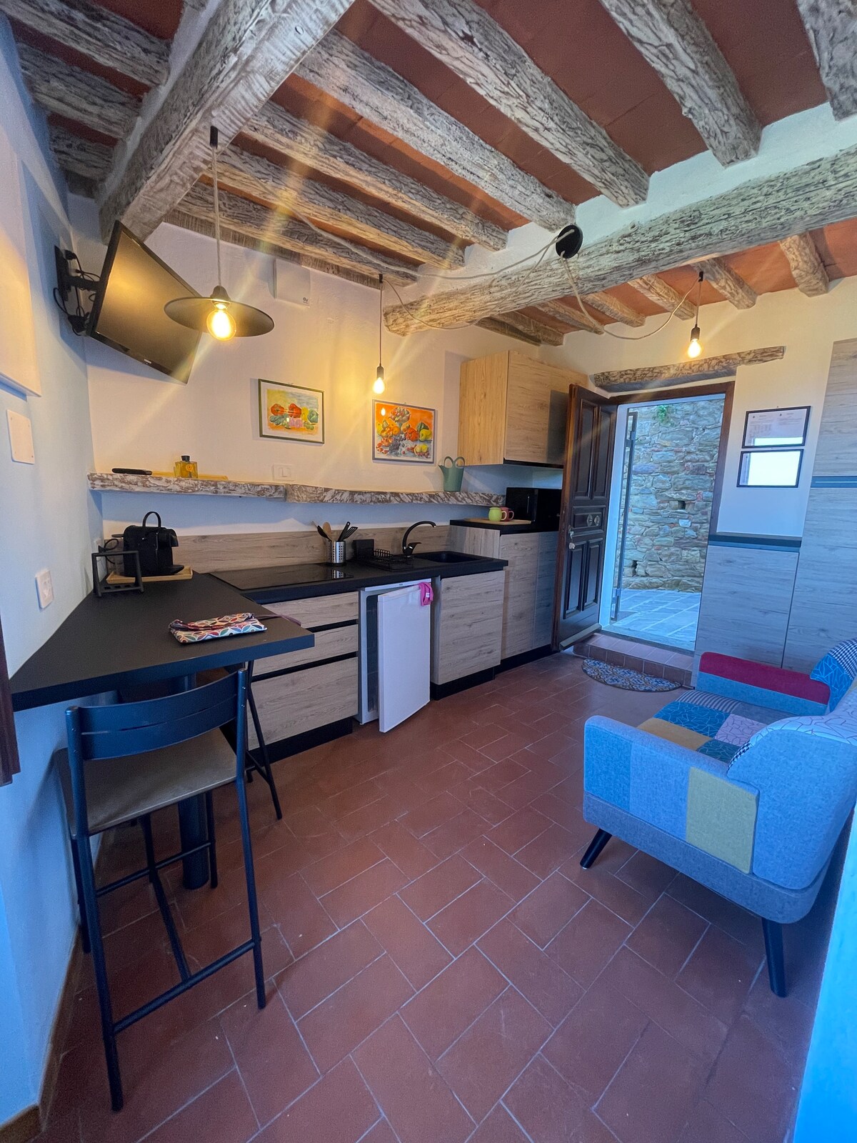 A compact kitchen area is equipped with modern appliances, including a refrigerator and a coffee maker. Exposed wooden beams add character to the ceiling. An inviting seating area features a colorful chair and a small table, while a door leads to an outdoor space.