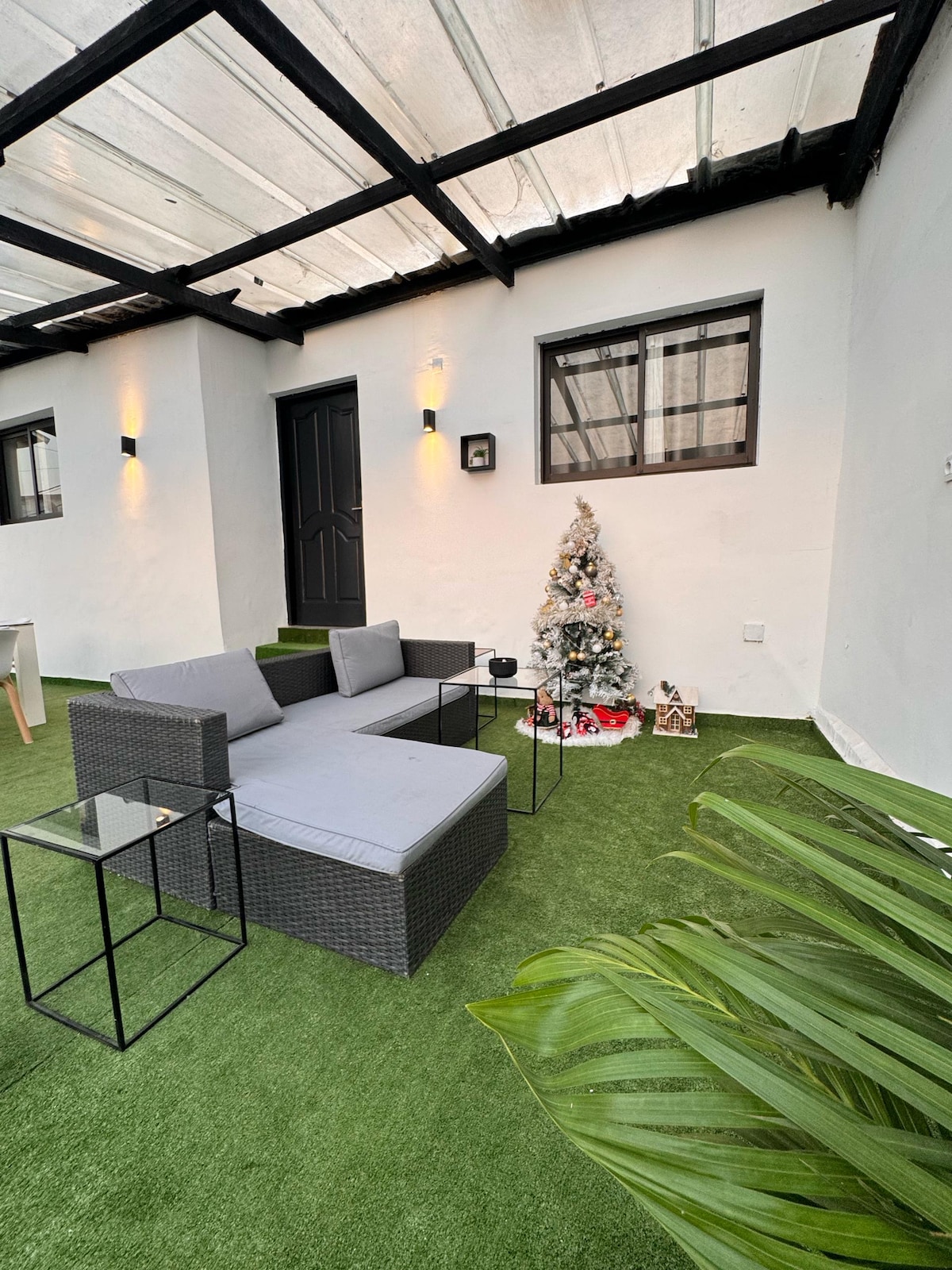 An outdoor seating area features a modern grey sectional sofa paired with two small black tables. Lush green artificial grass covers the ground, while a small Christmas tree adorned with festive decorations is positioned beside the sofa. Soft lighting enhances the inviting ambiance.
