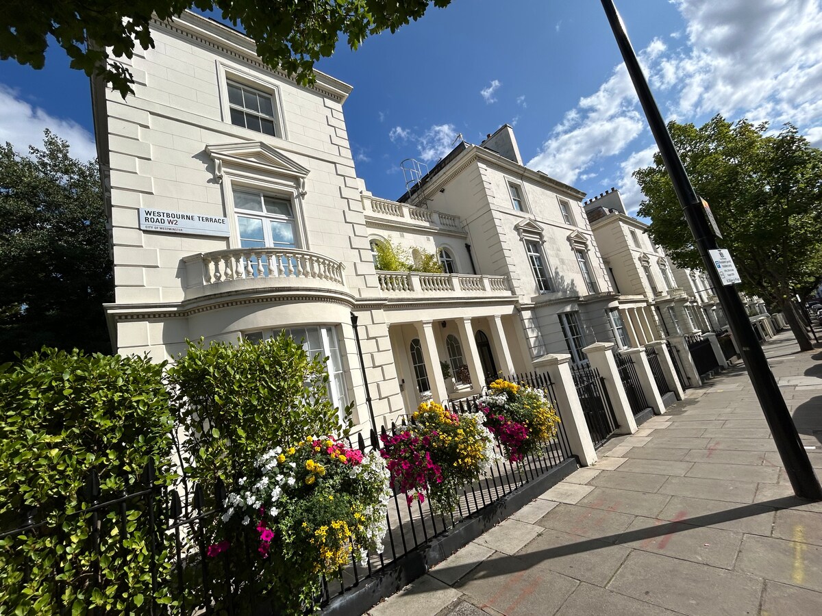A charming row of elegant white townhouses is showcased, featuring classic architectural details. Lush green foliage and colorful flower beds are visible in front of the properties, adding a touch of vibrancy to the scenic street lined with trees under a bright blue sky.