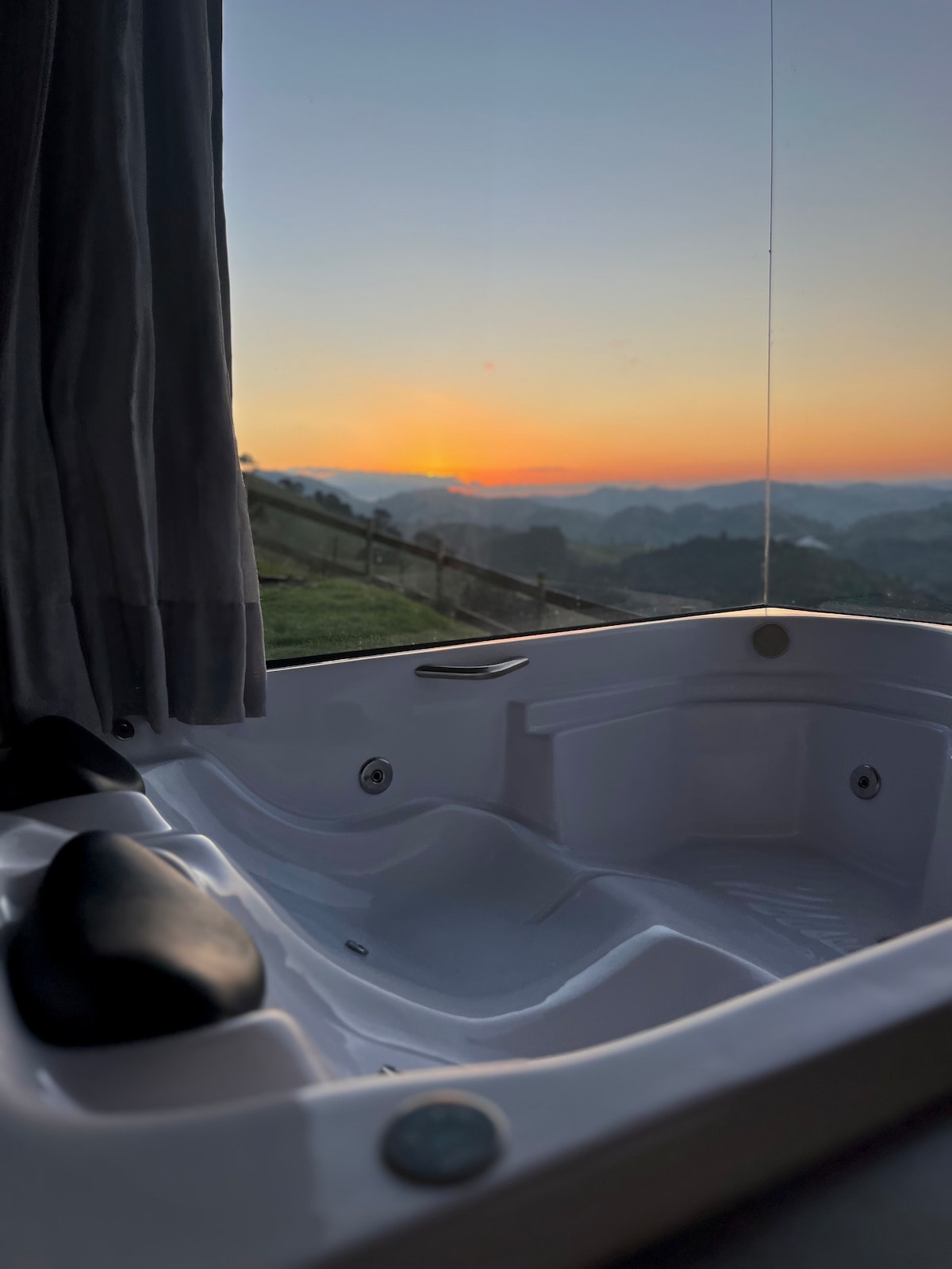 A hot tub is positioned by a large window, overlooking a scenic sunset. Soft light filters through sheer curtains, illuminating the tub's smooth surfaces. The distant mountains create a serene backdrop, suggesting a peaceful retreat into nature.