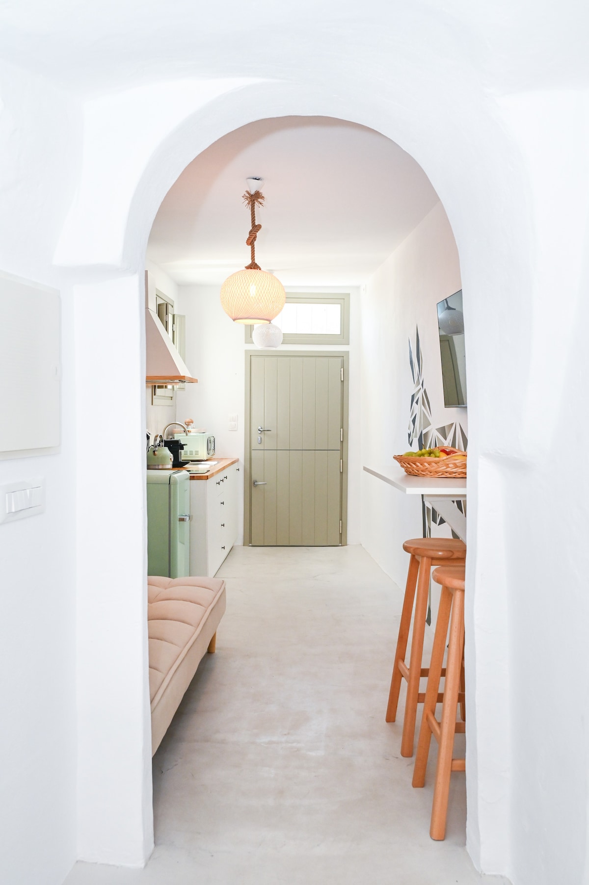 A bright hallway leads into the kitchen area, showcasing a light color palette. A wooden bar with two stools is positioned against the wall, complemented by a decorative bowl. An arched entrance adds architectural interest and softens the overall space.