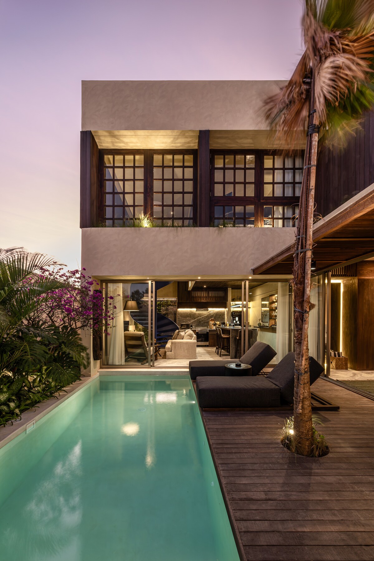 The two-story villa showcases a modern architectural design with large glass windows. A private pool is framed by wooden decking and tropical plants, creating a serene outdoor space. Lounge chairs are positioned beside the pool, providing a relaxing area to unwind.