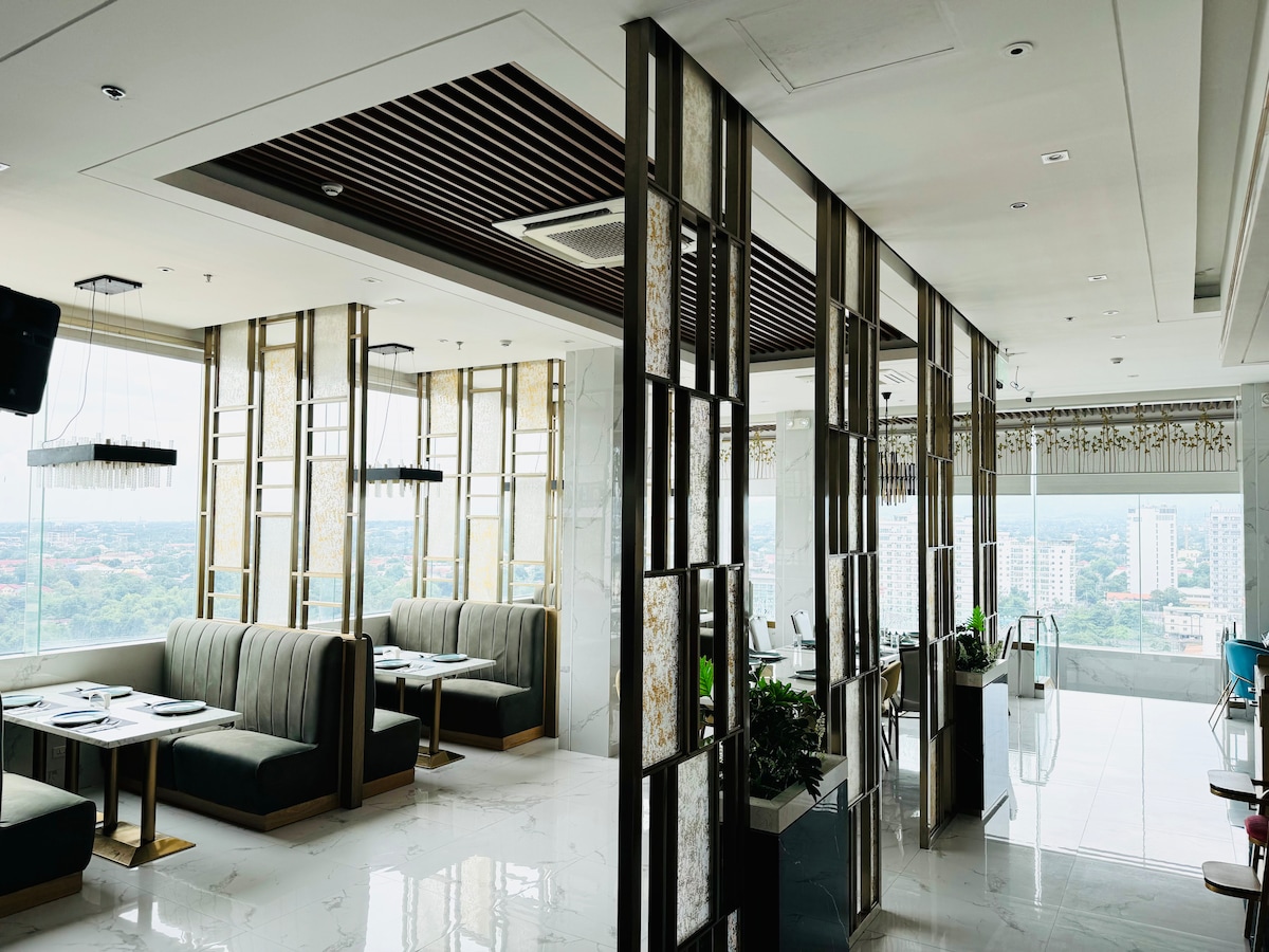 An elegant dining area features plush green banquettes and contemporary lighting. Floor-to-ceiling windows provide expansive views of the cityscape. A sleek partition adds a modern touch while allowing for an airy feel. The marble floors reflect the natural light, creating a bright atmosphere.