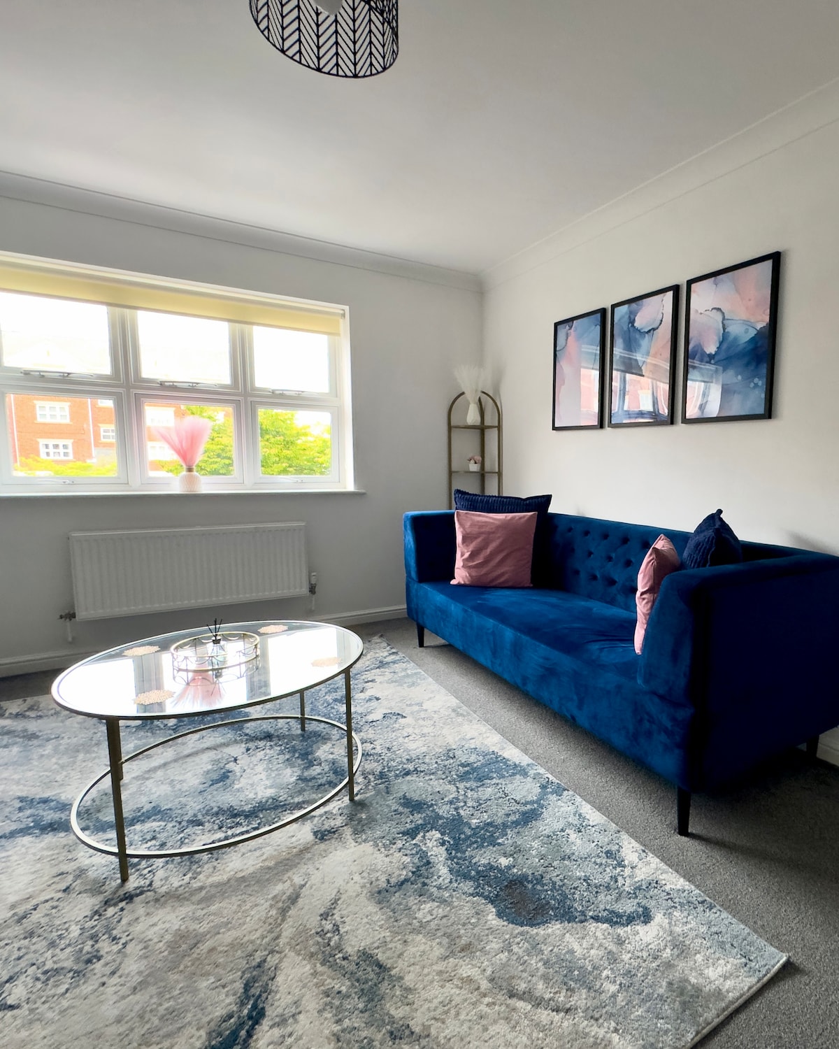 A modern living area features a blue upholstered sofa with decorative pillows. A glass coffee table sits on a patterned rug, while large windows provide natural light and a view of the surroundings. Artwork in soft hues adorns the wall, enhancing the space.