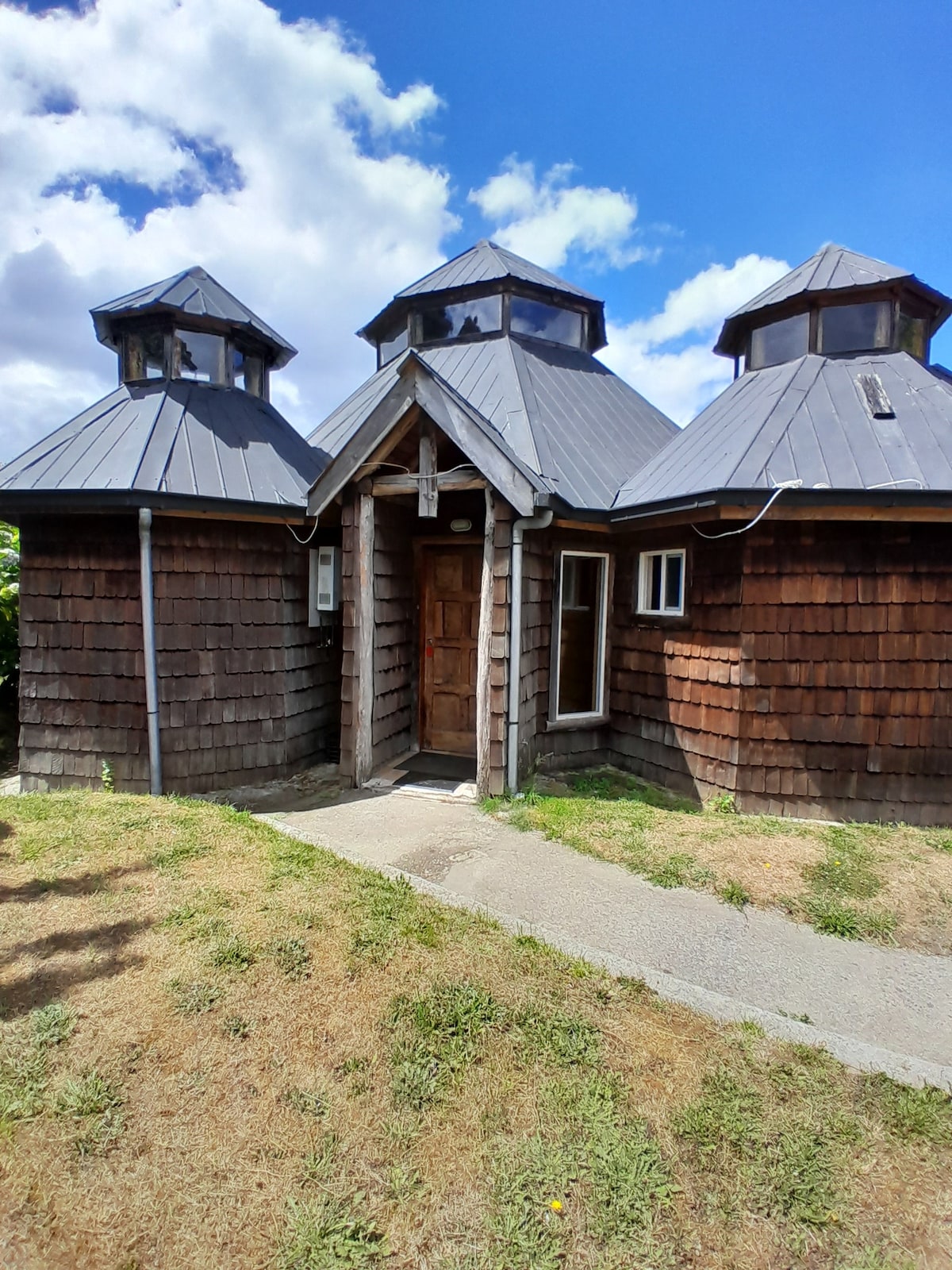 A unique wooden structure with three distinct peaks is shown, each featuring a conical roof. The entrance is framed by wooden beams, and a small window is visible on one side. The pathway is bordered by green grass and appears well-maintained.