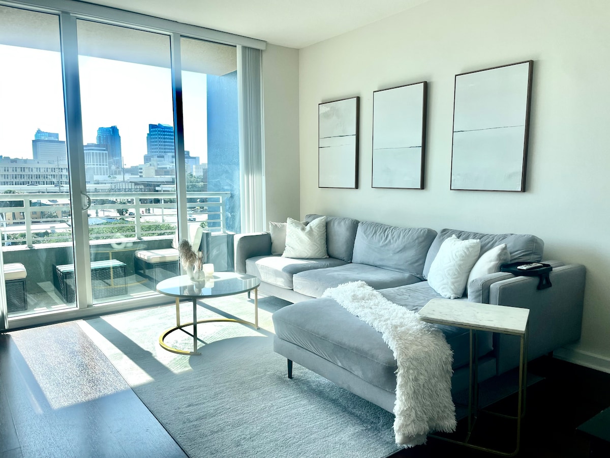 A modern living area features a light gray sectional sofa with an accent throw. A glass coffee table is positioned on a soft area rug, reflecting natural light from the floor-to-ceiling windows that offer views of the cityscape.