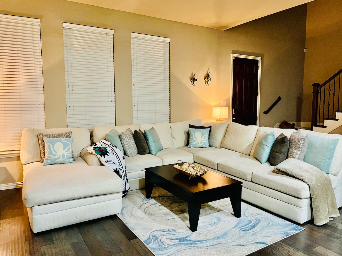 A spacious sectional sofa, adorned with decorative pillows in various shades, is positioned in a cozy living area. A coffee table with a decorative centerpiece rests on an area rug, while two large windows allow soft light to filter through the blinds.