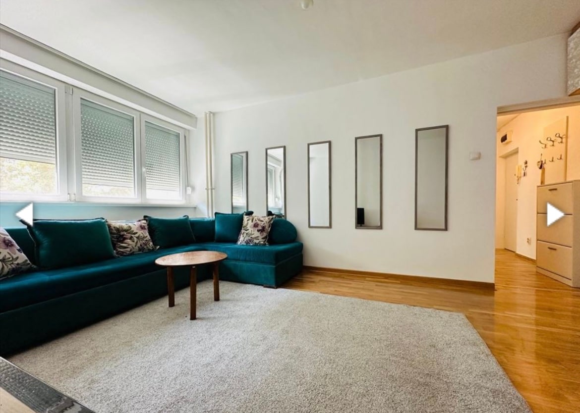 A spacious living area is featured, with a turquoise sectional sofa adorned with decorative pillows. Natural light enters through large windows, highlighting a neutral-toned rug on the wooden flooring. A small round table sits at the center, complemented by a wall of mirrors reflecting the space.