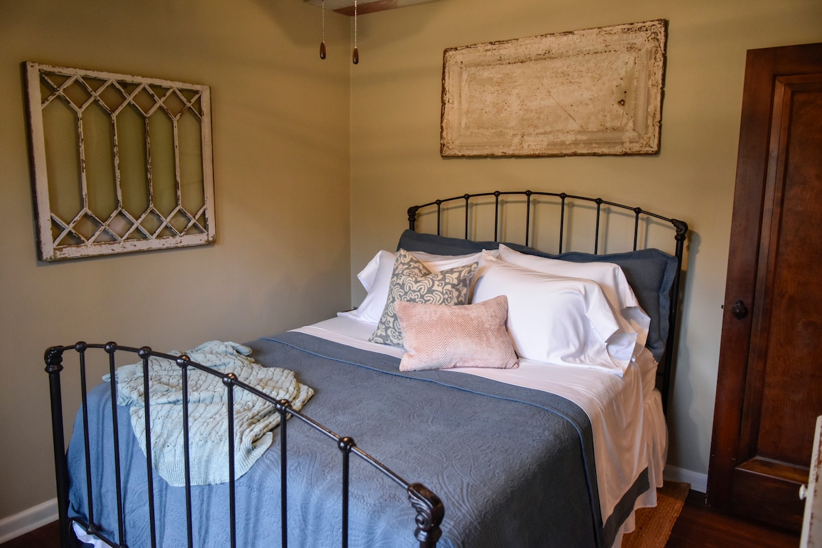 This charming second bedroom blends vintage farmhouse character with modern comfort. Featuring a cozy iron bed dressed in crisp linens, reclaimed wood ceilings, and curated antique décor, it’s the perfect quiet retreat after a day exploring the farm.